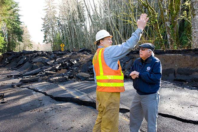 This photo shows the disastrous effects of flood damage on a road. With the broken road piled up in the background two men stand in the foreground of the photo discussing the natural disaster that occurred.